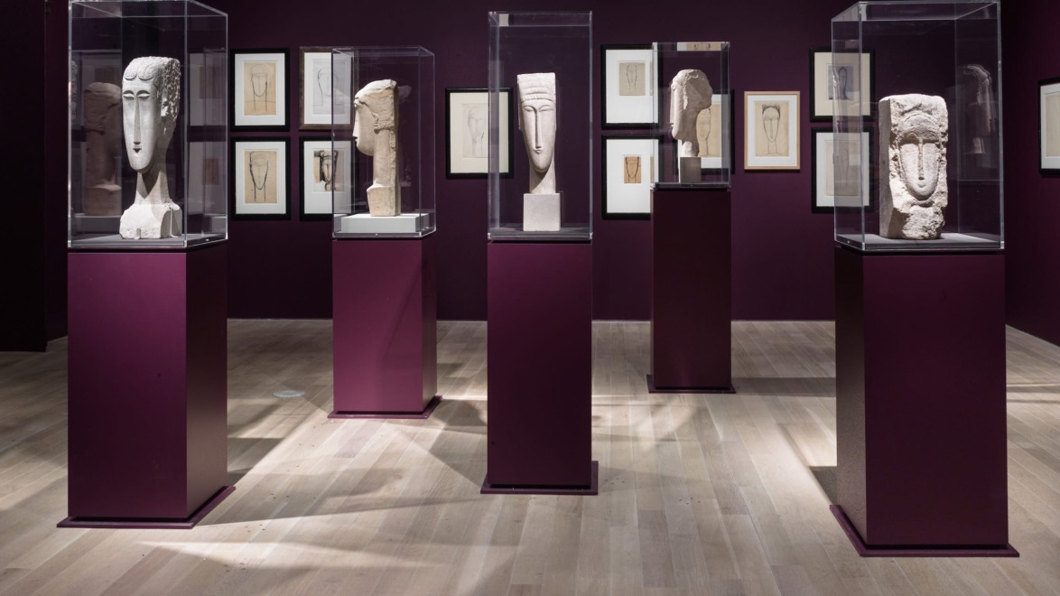 Exhibition room displaying several sculpted head busts in glass cases on burgundy pedestals with framed drawings on the walls.