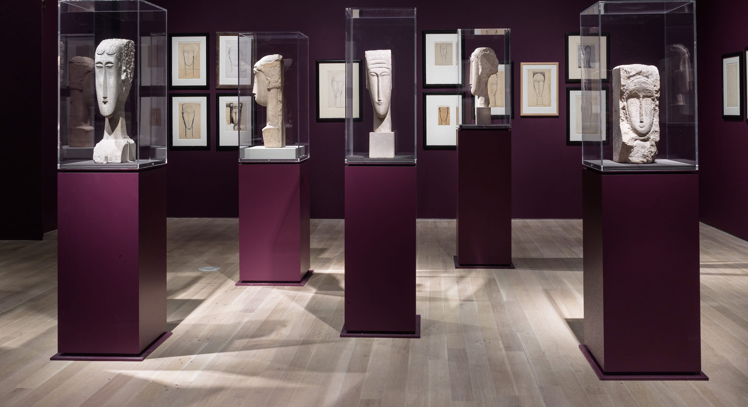 Exhibition room displaying several sculpted head busts in glass cases on burgundy pedestals with framed drawings on the walls.