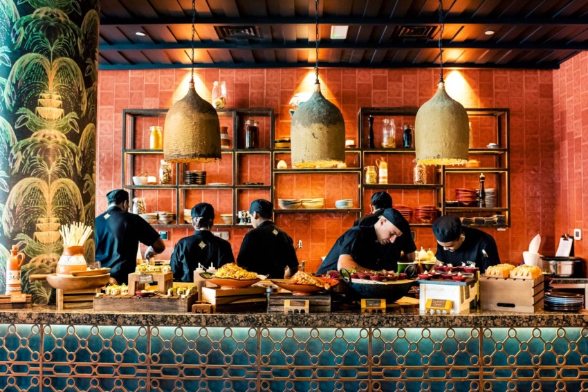 Chefs in a restaurant kitchen preparing various dishes with decorative lamps and an array of food ingredients on the counter.