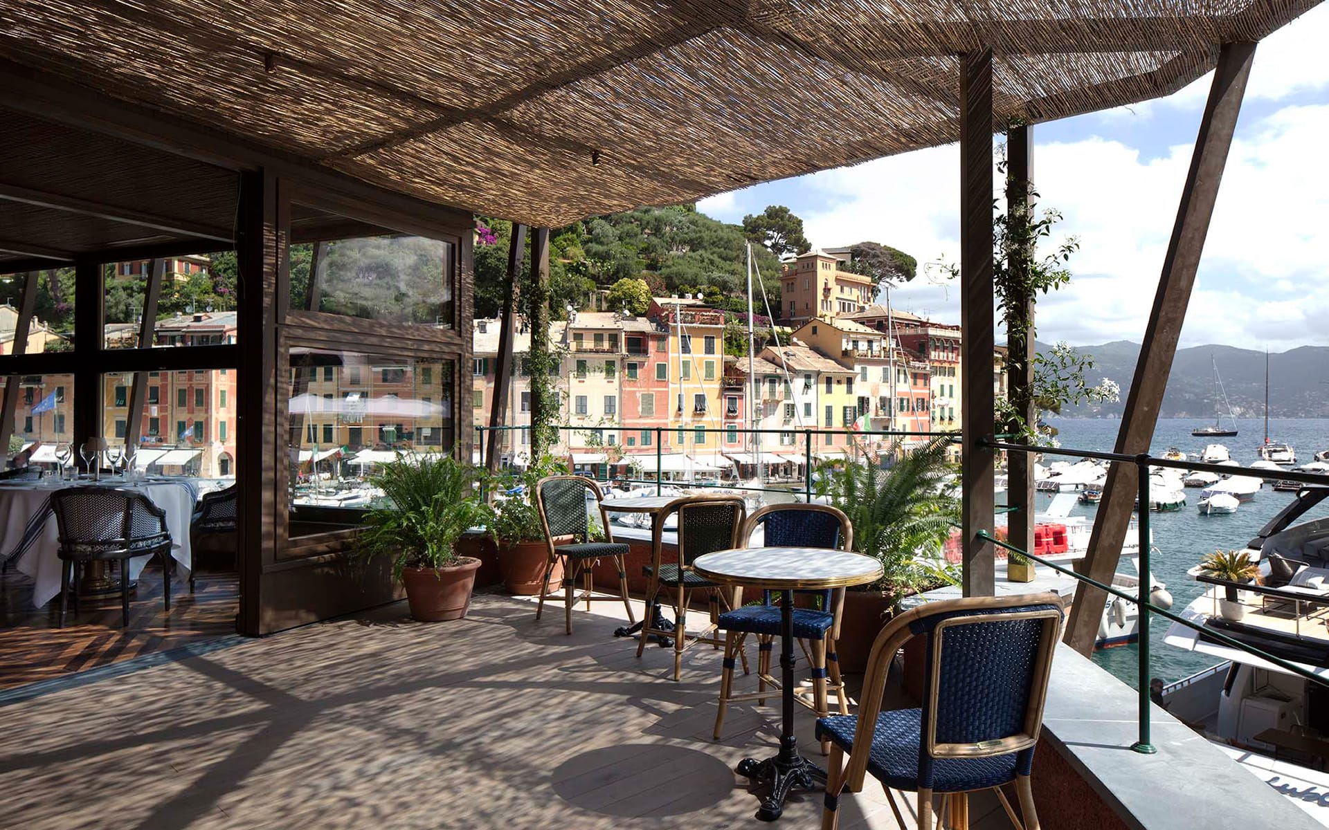 Outdoor terrace with tables and chairs overlooking a marina, colorful buildings, and lush greenery under a clear sky.