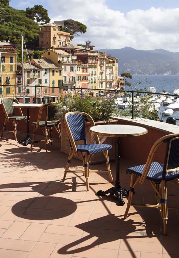 Outdoor café with empty chairs and tables overlooking colorful buildings and boats in a sunny coastal town.