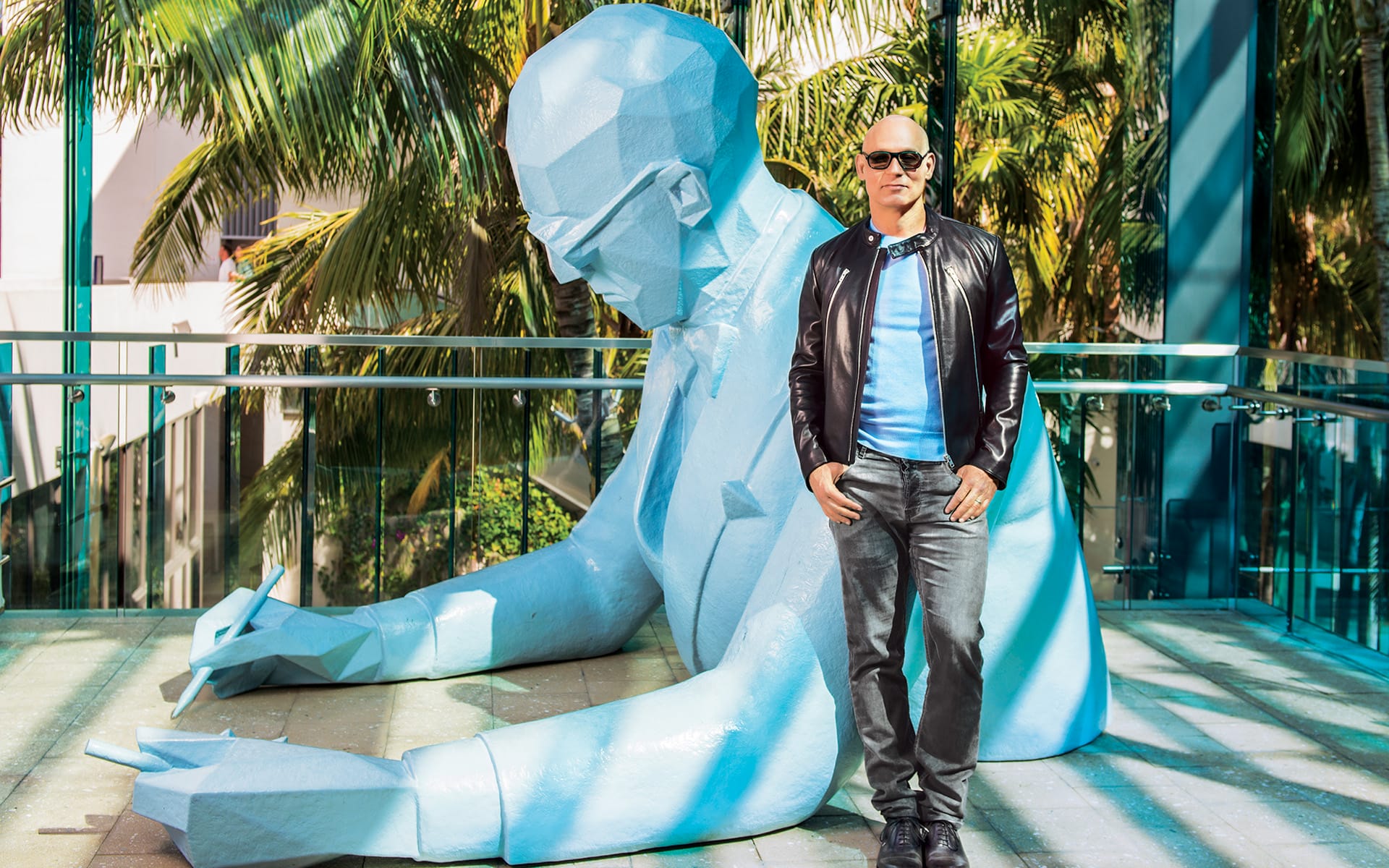 Man in leather jacket stands beside large blue sculpture of a person writing, with palm trees in the background.