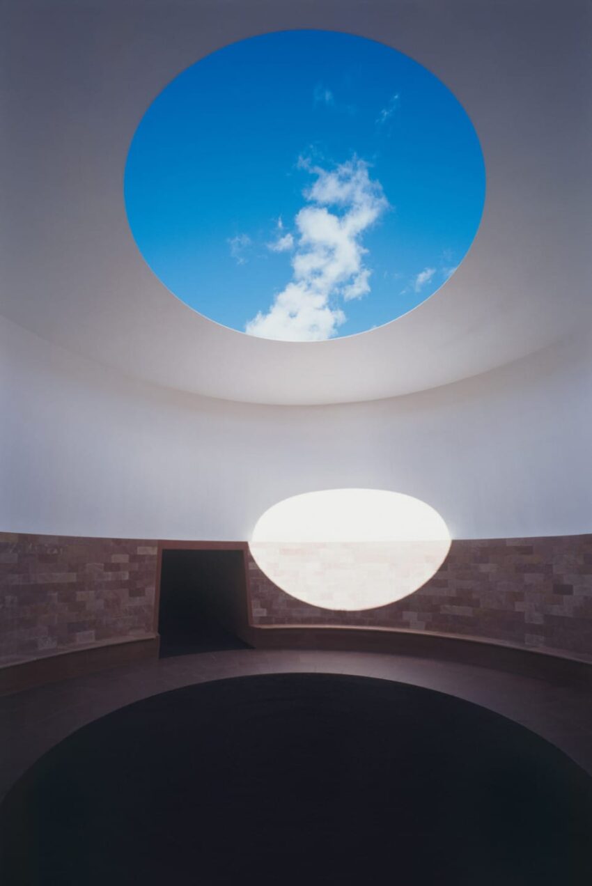 Circular skylight in a minimalist room with a view of blue sky and clouds above a stone wall and dark floor.