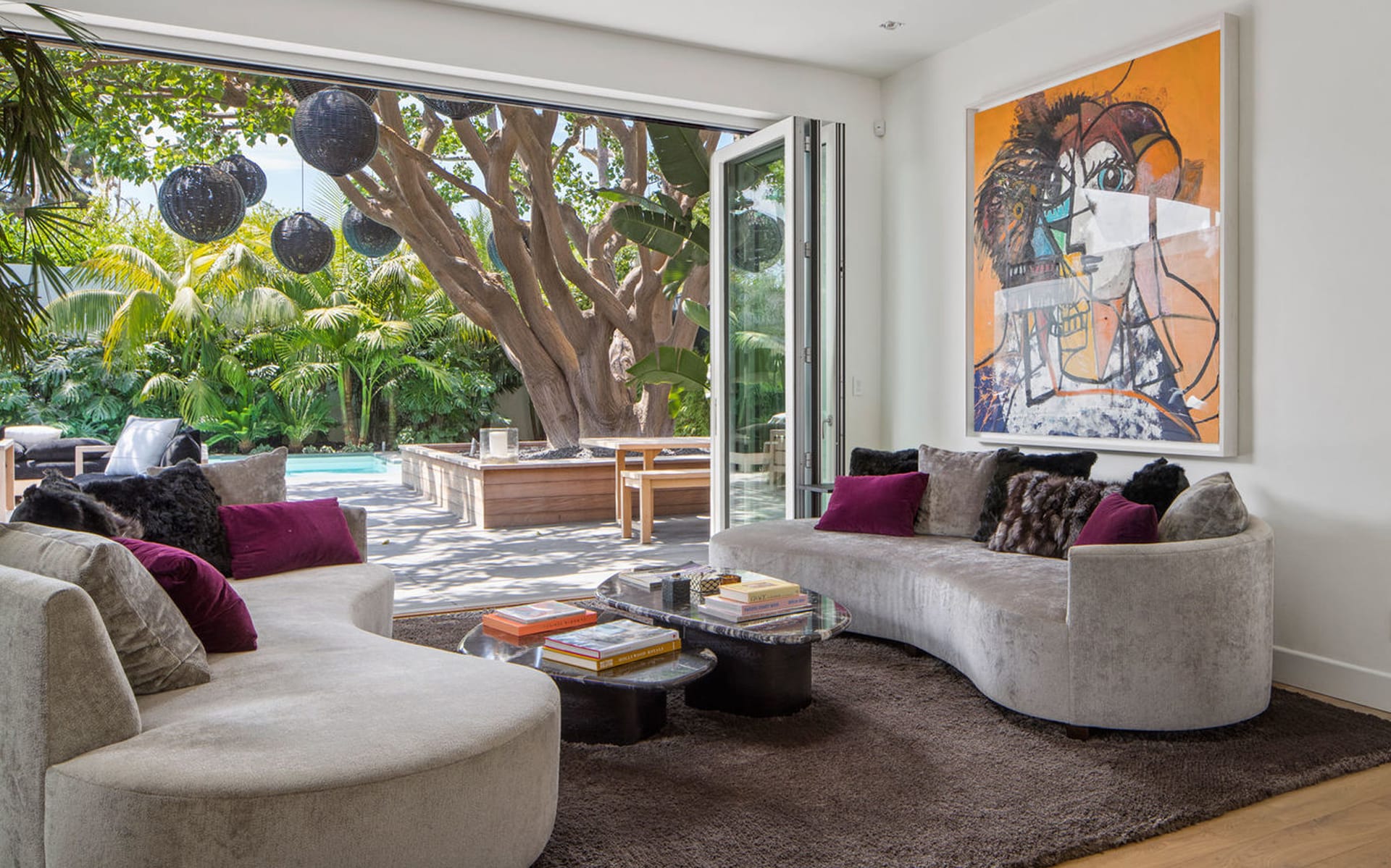 Modern living room with curved sofas, abstract art, and open glass doors revealing lush garden and pool area.
