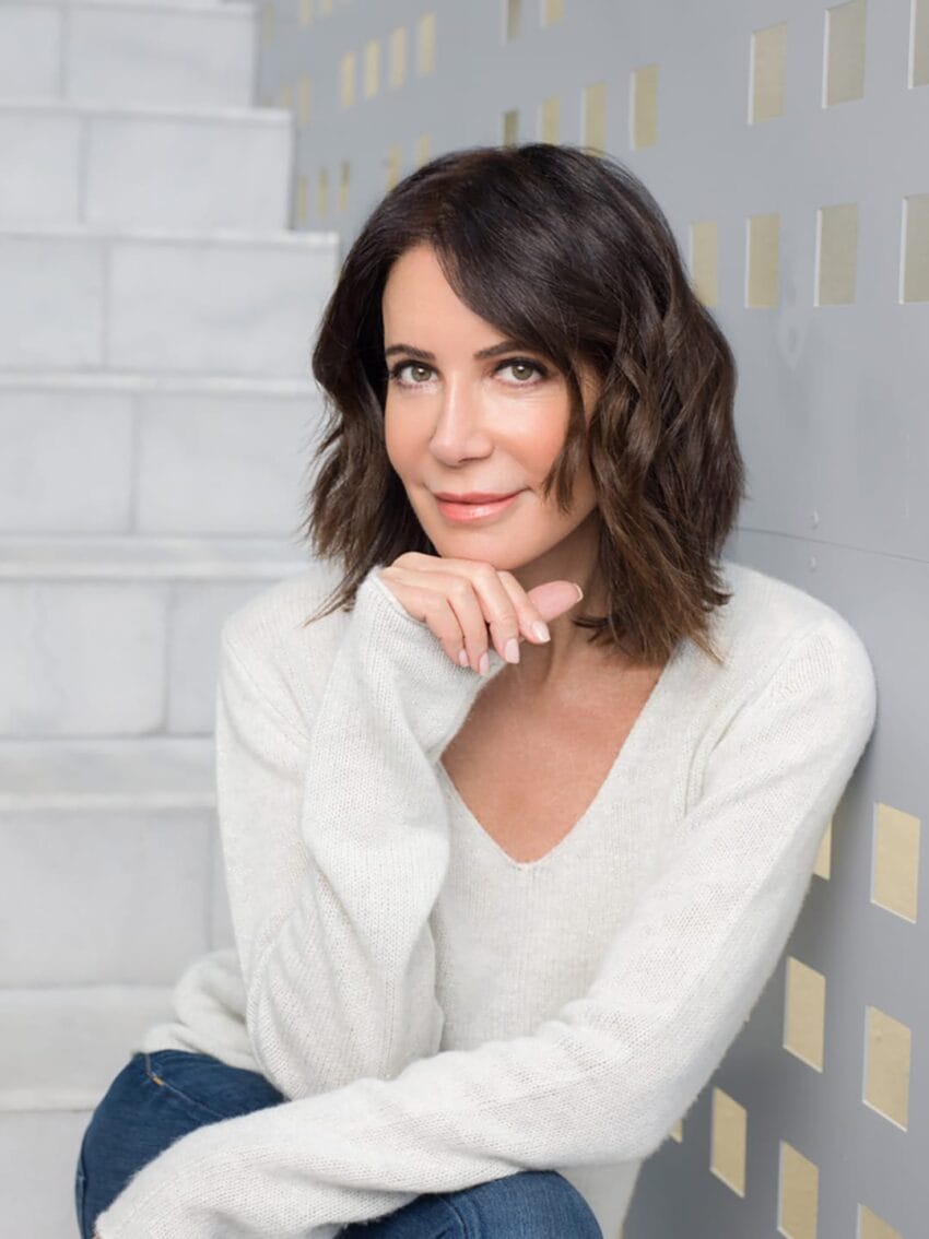 Person with short brown hair sitting on stairs, wearing a white sweater, smiling gently with hand resting on chin.