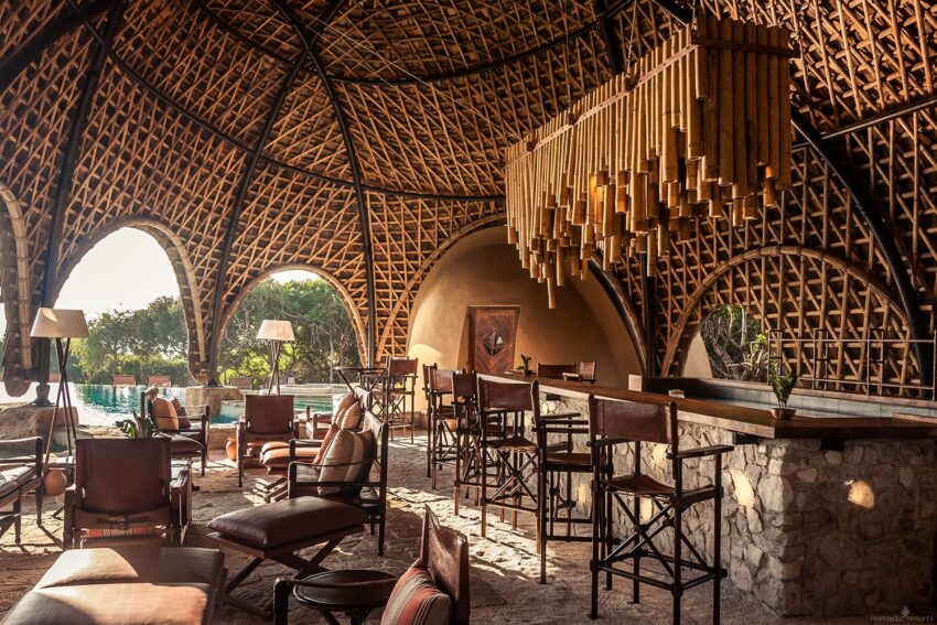 Luxurious tropical resort lounge with wooden chairs, a stone bar, and bamboo ceiling, overlooking a pool and lush greenery.