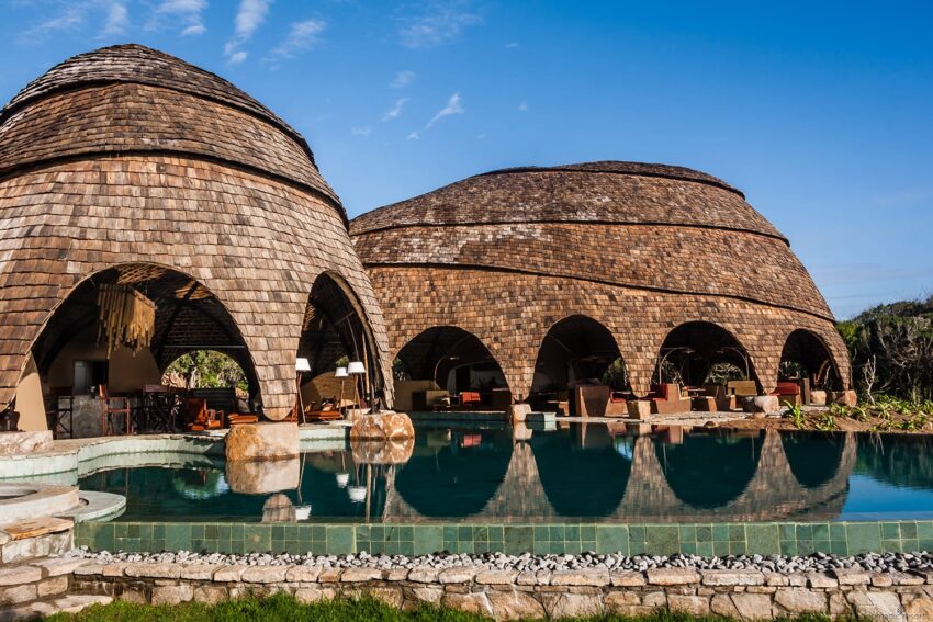 Unique domed buildings with wooden shingles beside a tranquil pool under a clear blue sky.