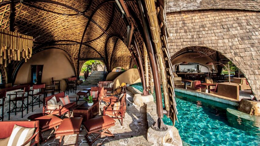 Outdoor restaurant with wooden architecture, poolside seating, cushioned chairs, and tables under a woven, arched roof.