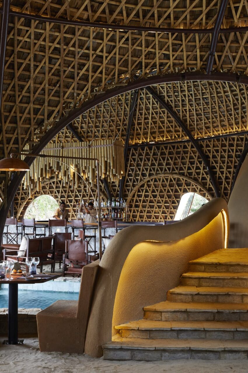 Interior view of a bamboo-structured cafe with a curved staircase, wooden ceiling, and seating area with visible dining table.