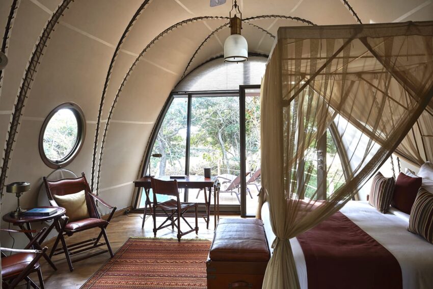 Luxurious safari lodge interior with arched ceiling, large window, cozy furniture, and canopy bed overlooking lush greenery.