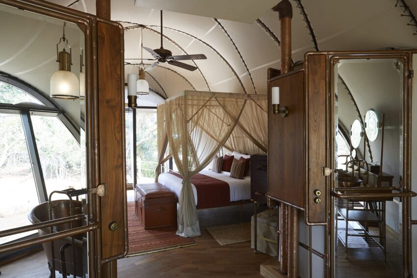Luxury safari lodge bedroom with canopy bed, wood and metal decor, and large windows overlooking a natural landscape.