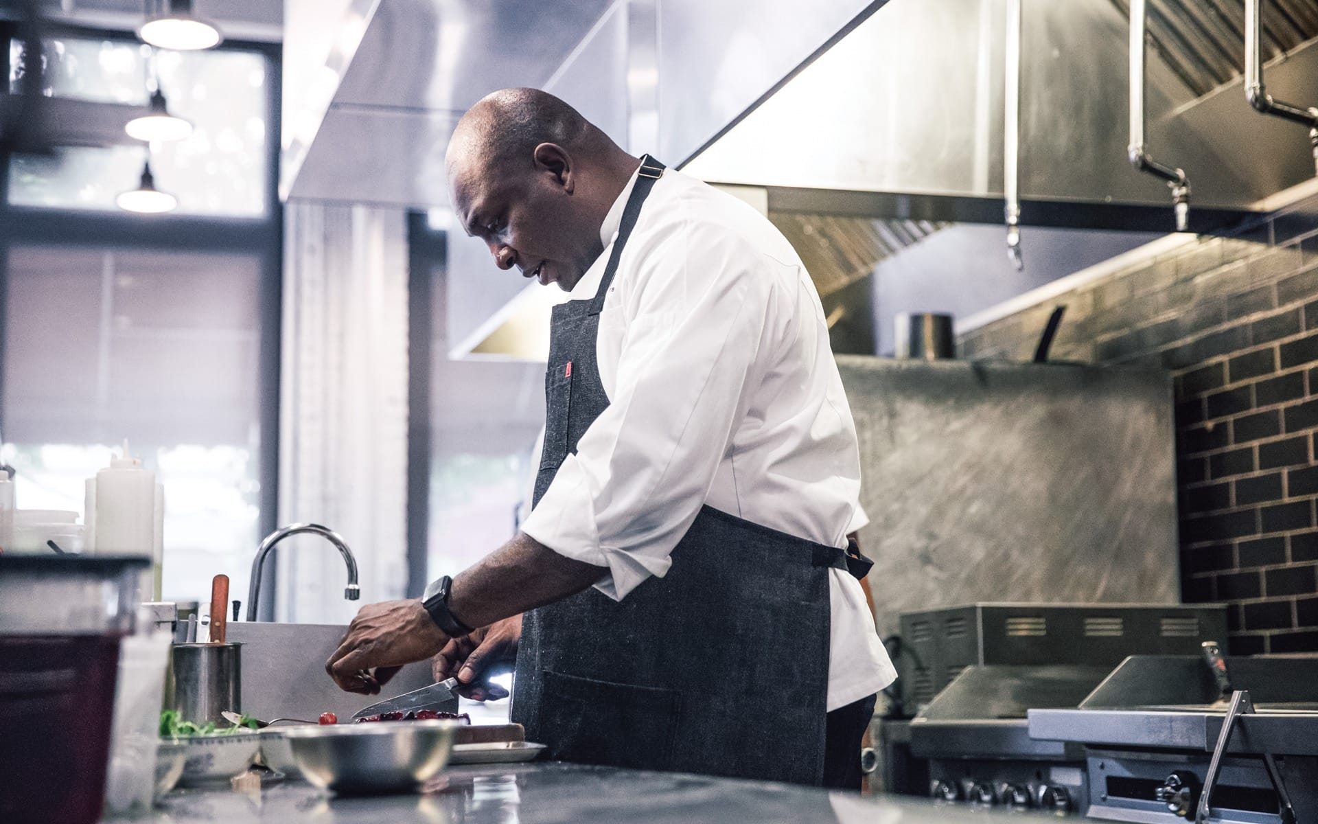 Chef in a commercial kitchen preparing food, wearing a white shirt and dark apron, focused on his task.