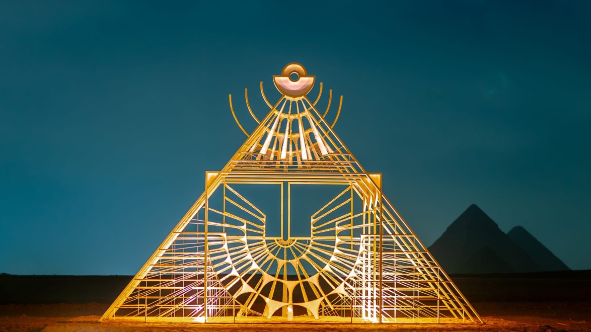 Illuminated geometric sculpture in front of pyramids at night with deep blue sky in the background