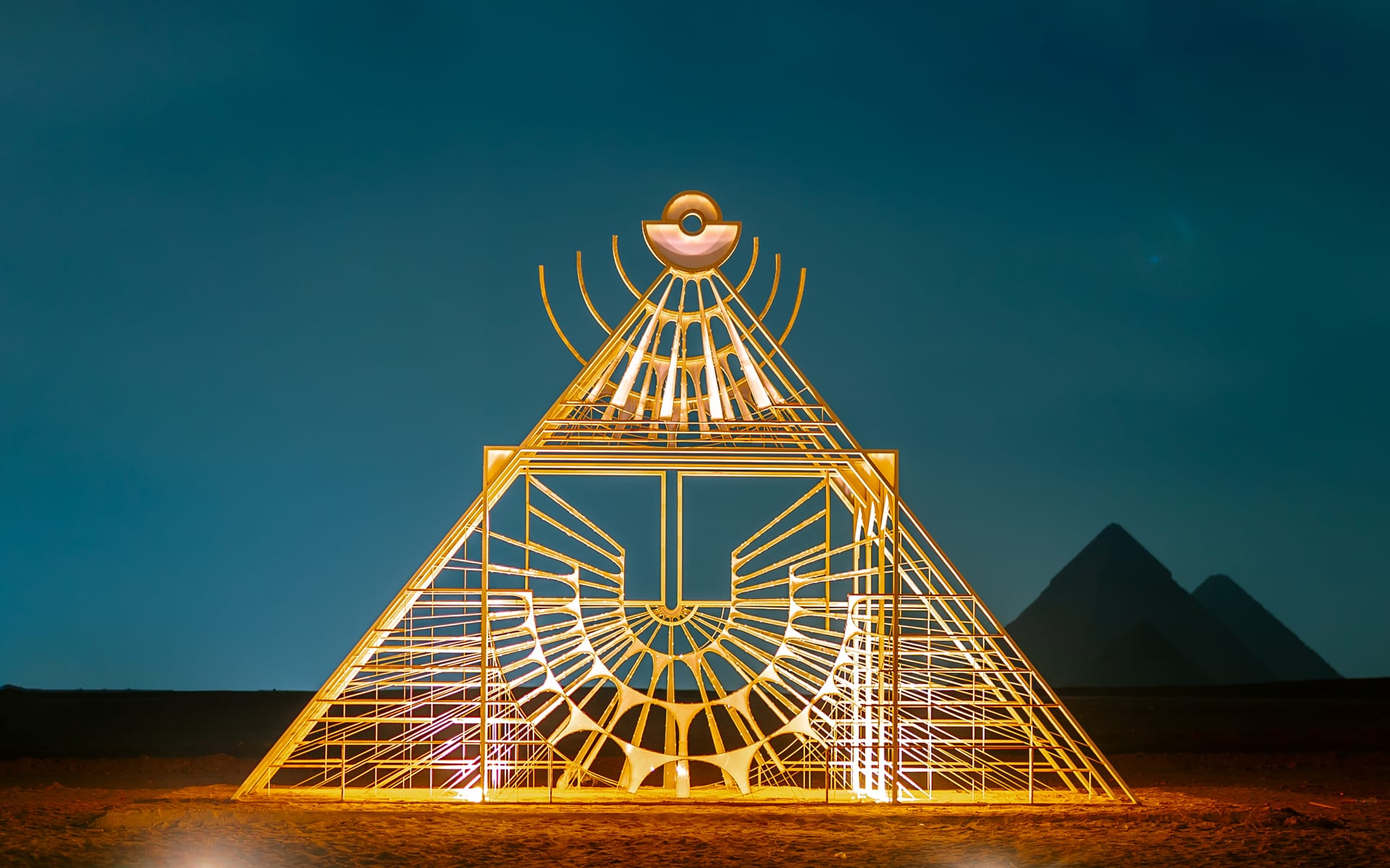 Illuminated geometric sculpture in front of pyramids at night with deep blue sky in the background