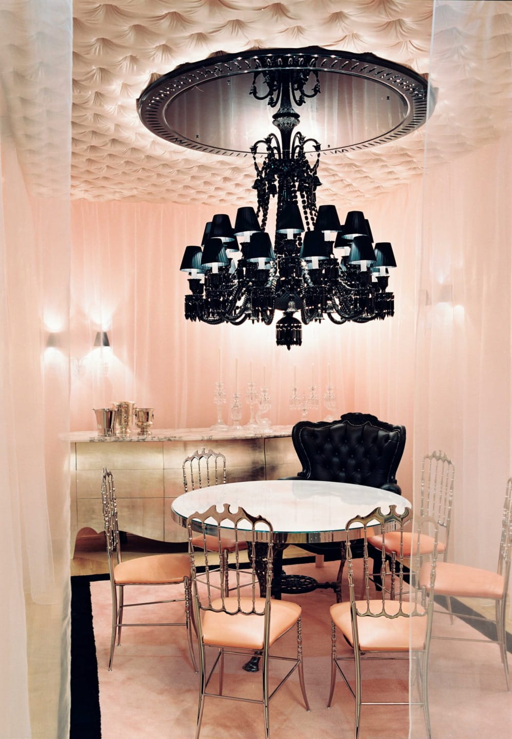 Elegant dining room with black chandelier, round table, silver chairs, tufted ceiling, and soft peach curtains.