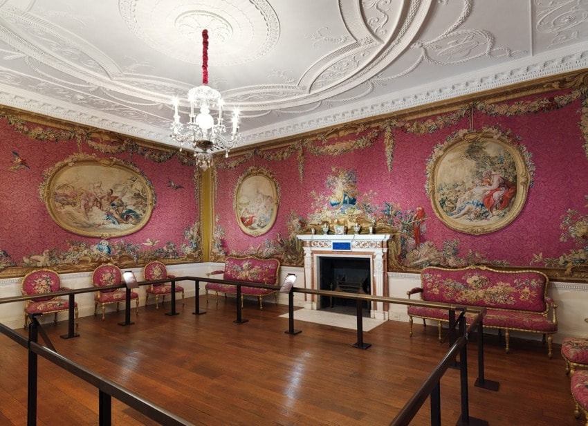 Elegant historical room with ornate red wallpaper, decorative ceiling, chandelier, fireplace, and classic furniture.