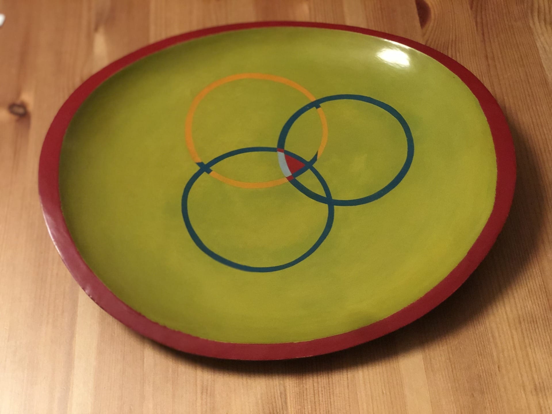 Green plate with overlapping red, blue, and orange circles design, red rim, on a wooden surface.
