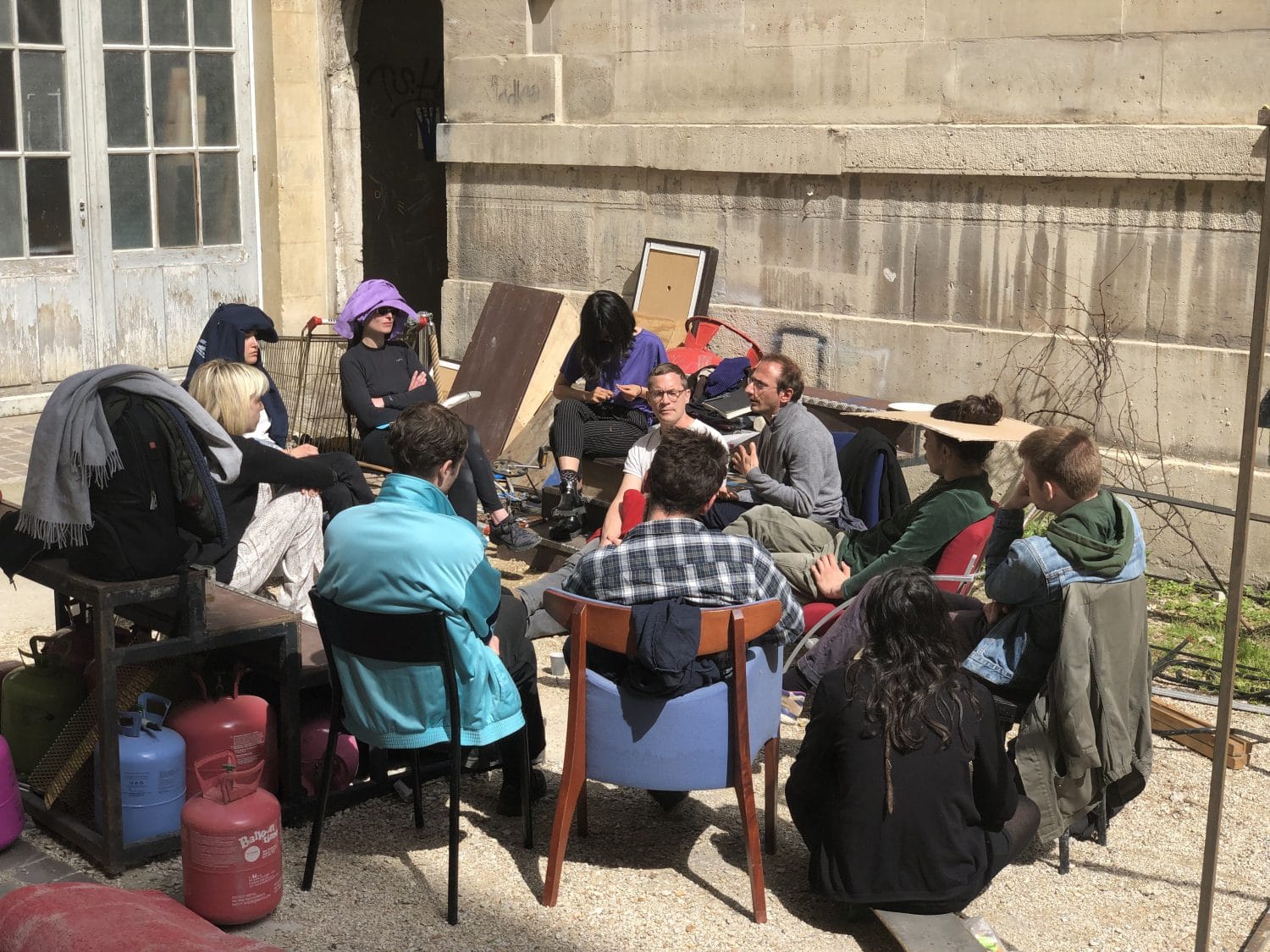 Group of people sitting in a circle outside, engaged in discussion near a building with wooden doors.