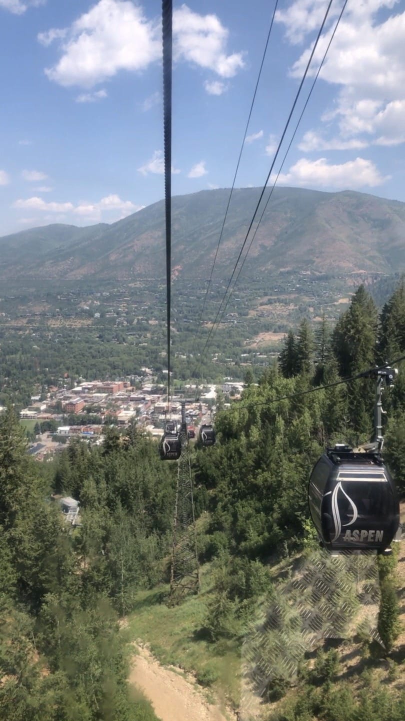 Gondola ride ascending over lush green forest with mountain and town view in the background under a partly cloudy sky.