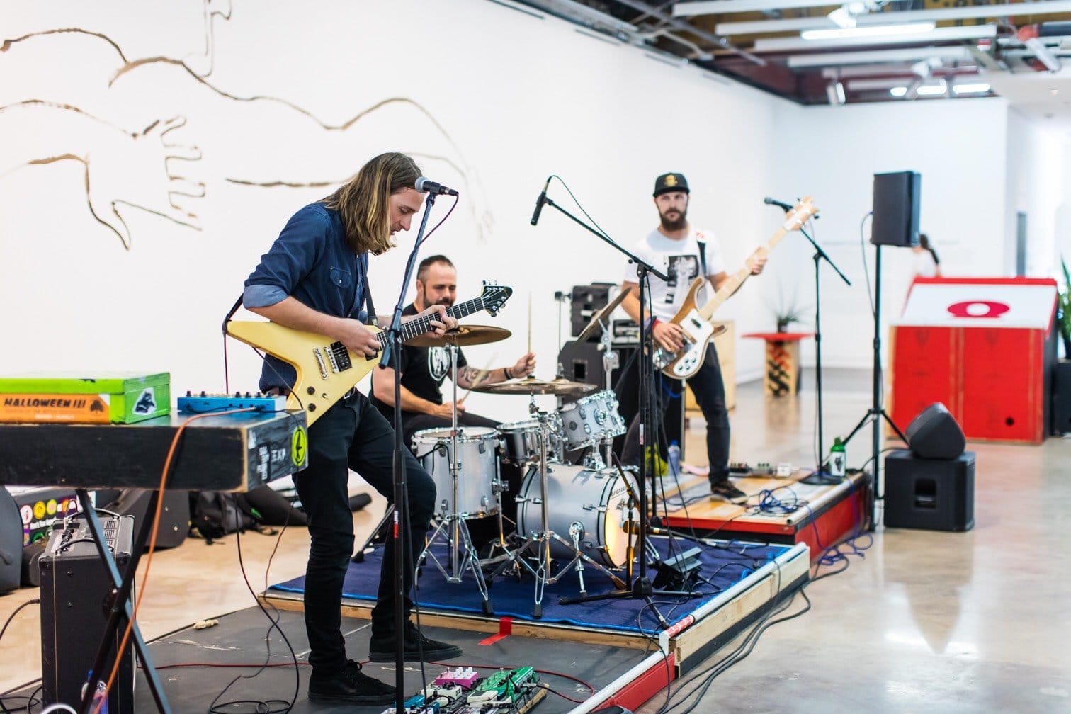 Three musicians perform in a modern art space with guitars and drums, surrounded by equipment and abstract wall art.