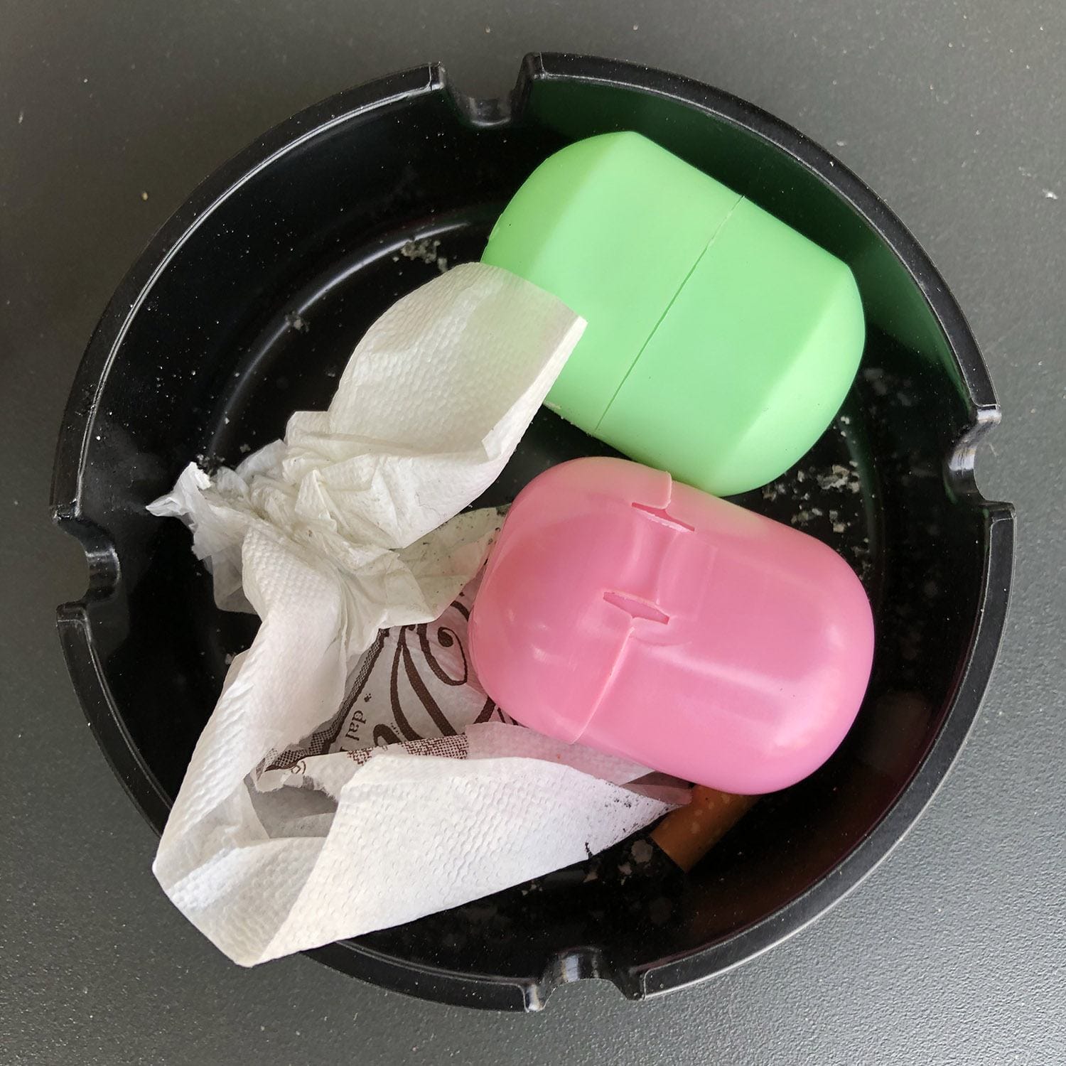 Pink and green plastic cases with tissues and a candy wrapper in a black ashtray.