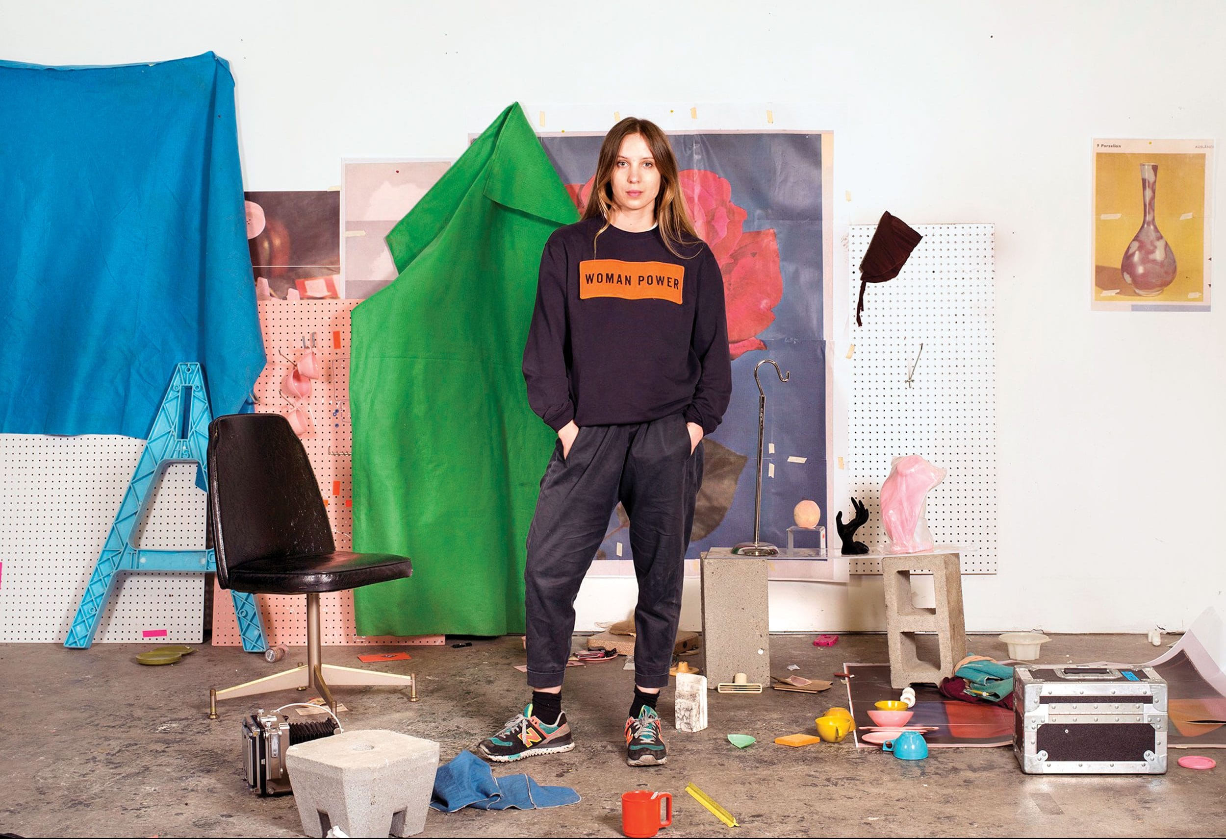 Person standing confidently in front of colorful backdrop with scattered items, wearing a sweatshirt that says "Woman Power".