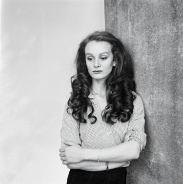 Woman with long curly hair standing against a concrete wall, arms crossed, wearing a checkered shirt. Black and white portrait.