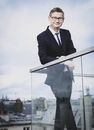 Man in a blue suit smiling while leaning on a glass railing with a cityscape in the background.