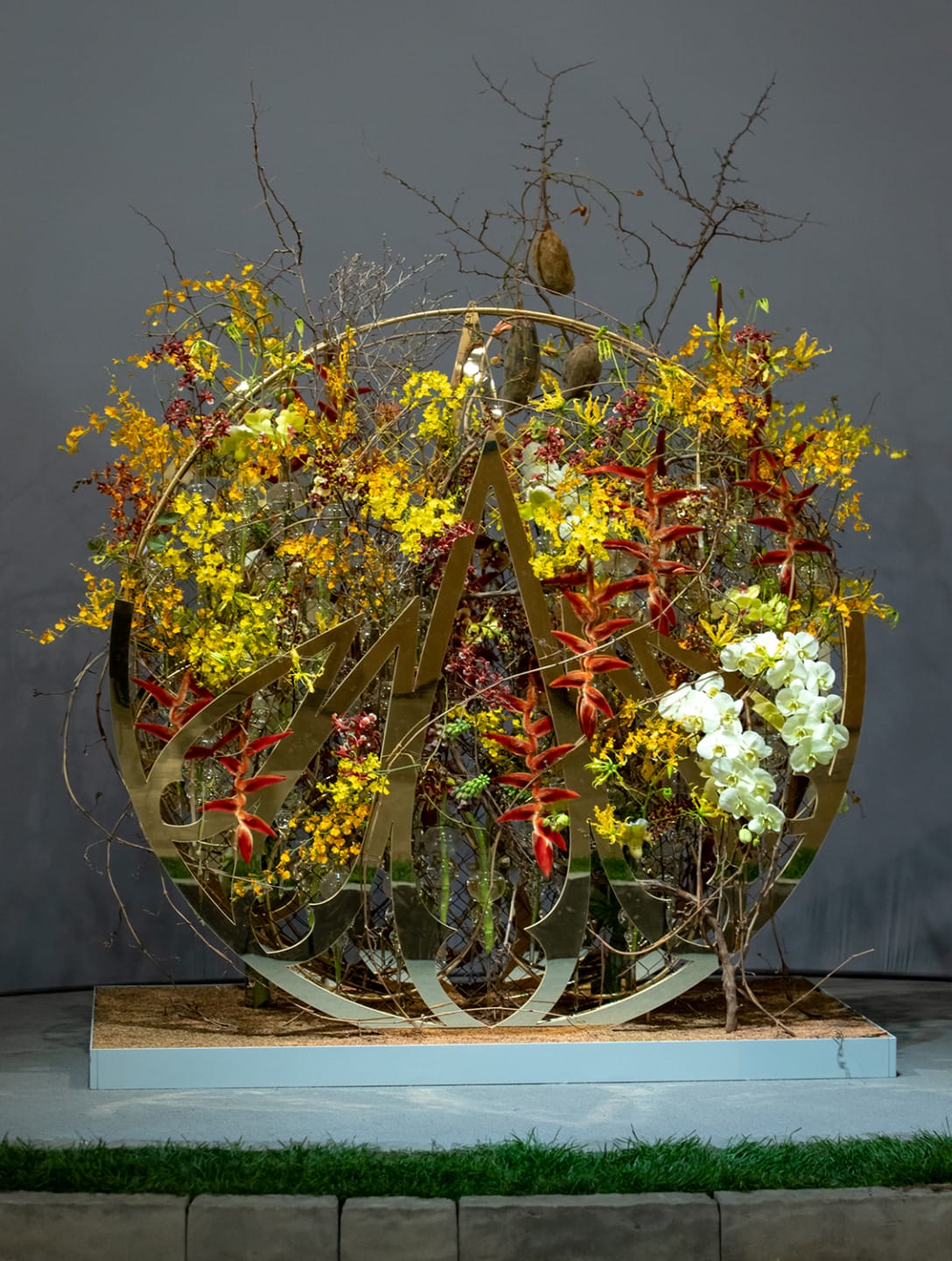 Elaborate floral display with yellow, red, and white flowers arranged on a metal structure, set against a gray background.