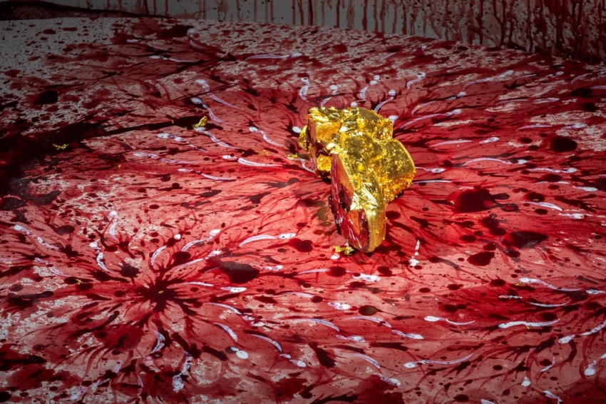 Abstract artwork with a vibrant red splatter pattern and a gold-painted rock placed at the center.