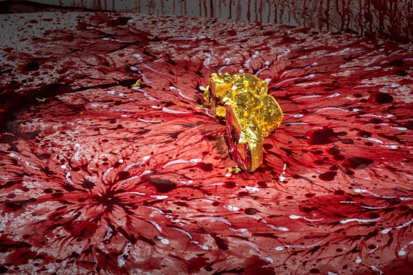 Abstract artwork with a vibrant red splatter pattern and a gold-painted rock placed at the center.
