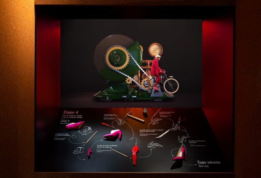 Window display with a vintage machine, man on a bicycle, step-by-step shoe process, and vibrant red high heels.