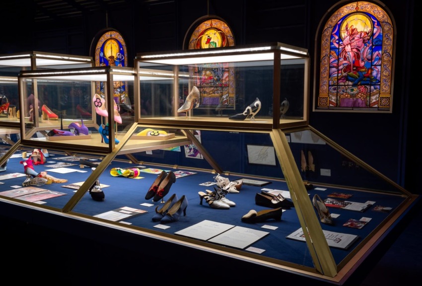 Exhibit showcasing various pairs of shoes displayed in glass cases with decorative stained glass windows in the background.