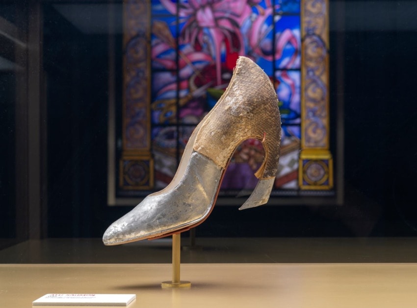 Silver and brown vintage shoe displayed in a glass case with an ornate stained glass background.