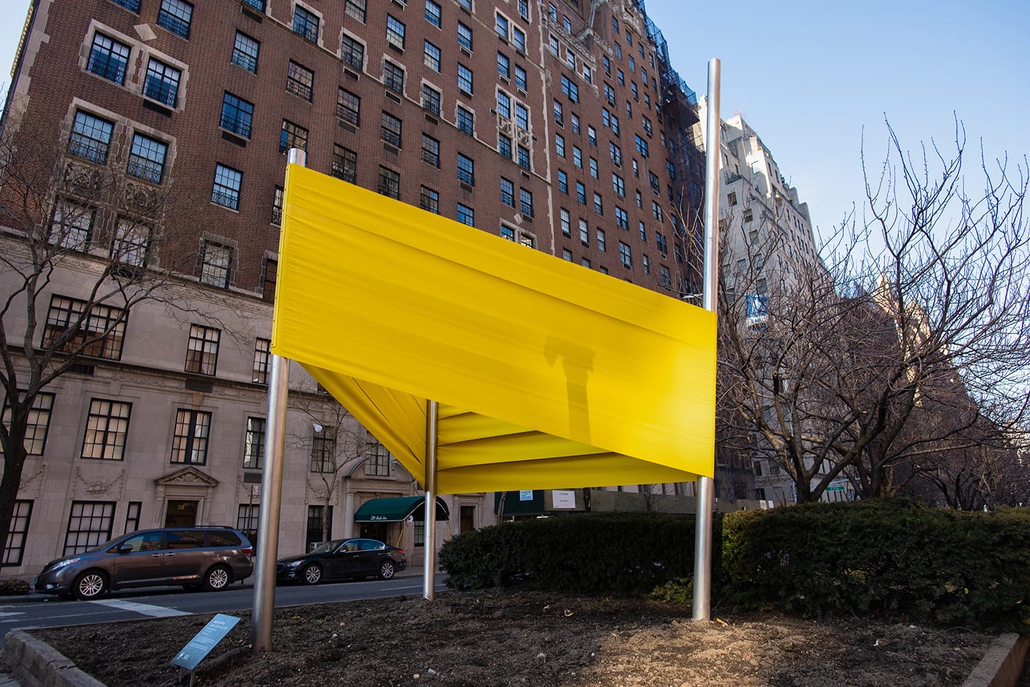 Yellow fabric installation in urban park, with apartment buildings and parked cars in the background on a clear day.