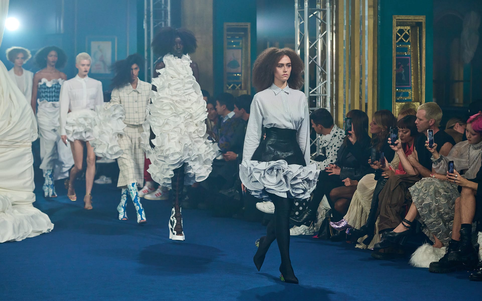 Fashion show with models wearing avant-garde outfits walking down a blue carpeted runway.