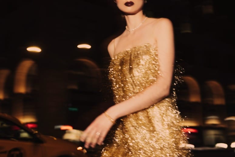 Person in shimmering gold dress walking on a city street at night with blurred lights in the background.