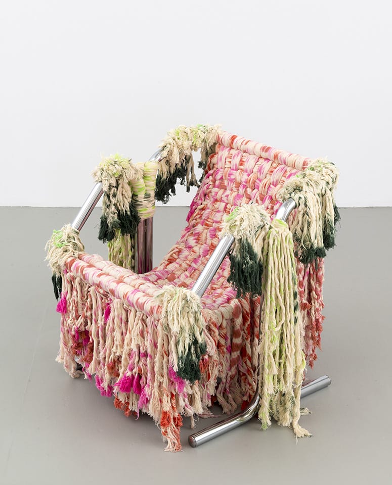 Colorful woven chair with fringe and metallic frame, featuring pink, green, and cream textures against a gray floor and white wall.