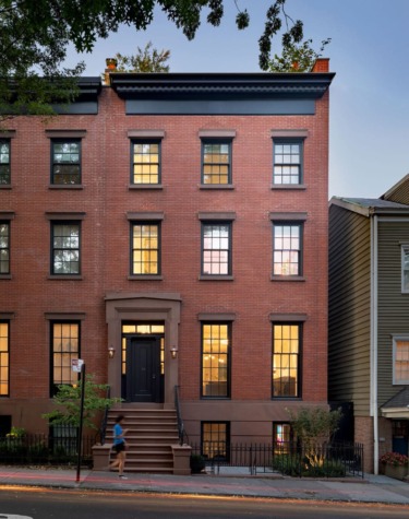 Three-story red brick townhouse with black trim, large windows, and a central entrance, at dusk with a person walking by.