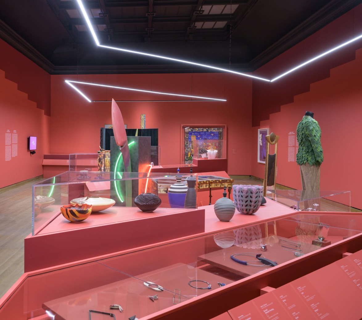 A museum exhibit with modern art pieces on display in a room with red walls and sleek ceiling lights.
