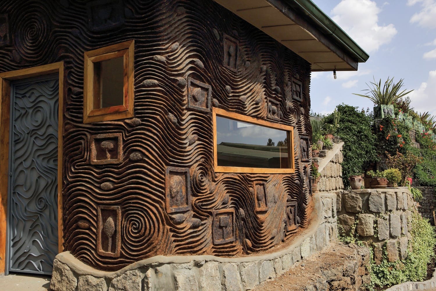 Eco-friendly house with intricate earthen wall design, featuring rectangular windows and surrounding lush greenery.