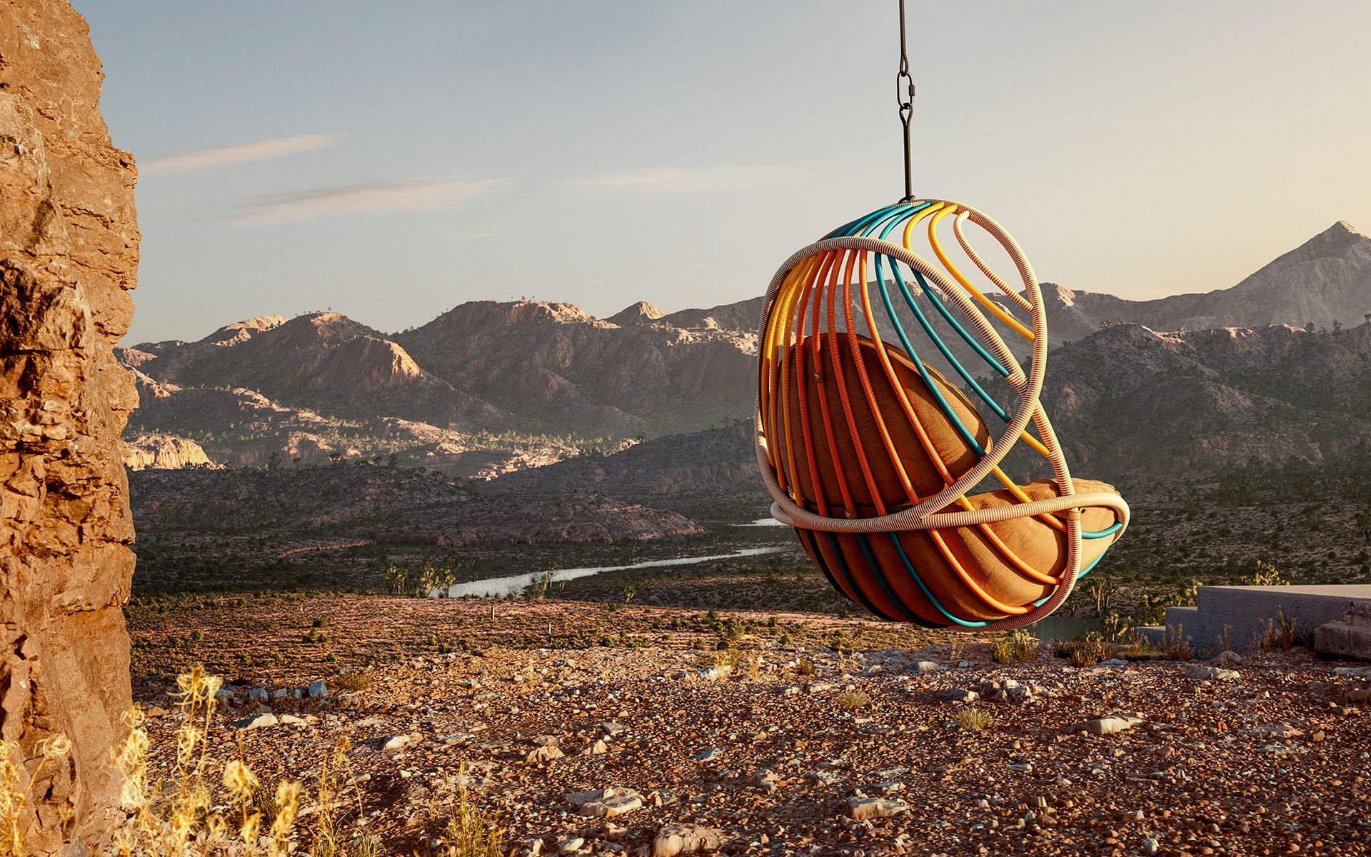 Hanging spherical chair with cushions overlooking a desert landscape with mountains and a river at sunset.