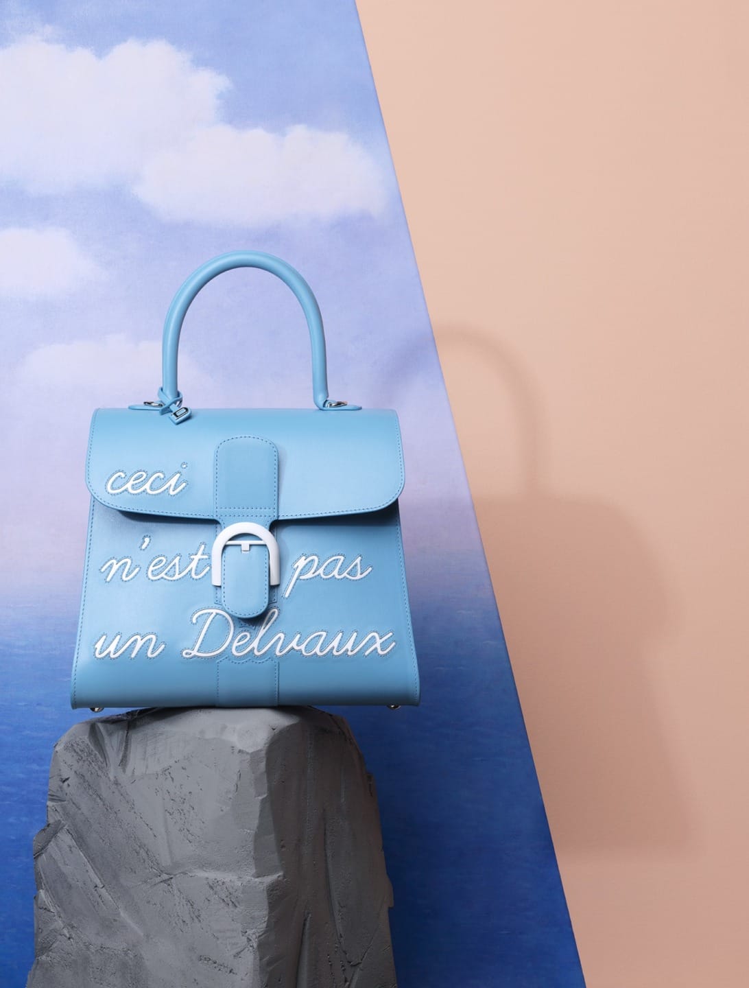 Blue handbag with French text on sea and sky background, positioned on a gray rock surface.