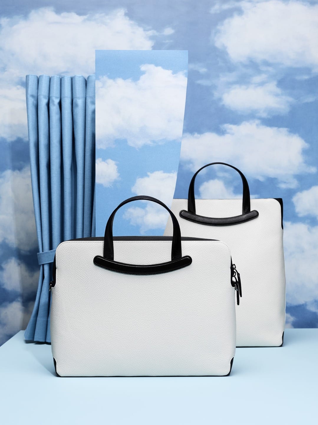Two white handbags with black handles in front of a sky-themed background with clouds and blue curtains.