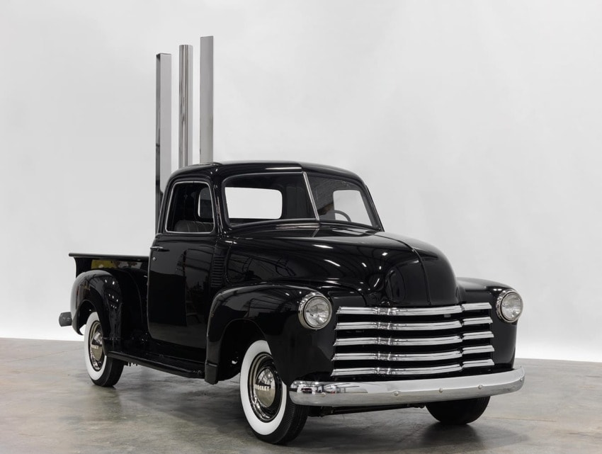Black vintage pickup truck with shiny chrome details and large exhaust stacks on display in a well-lit indoor setting.