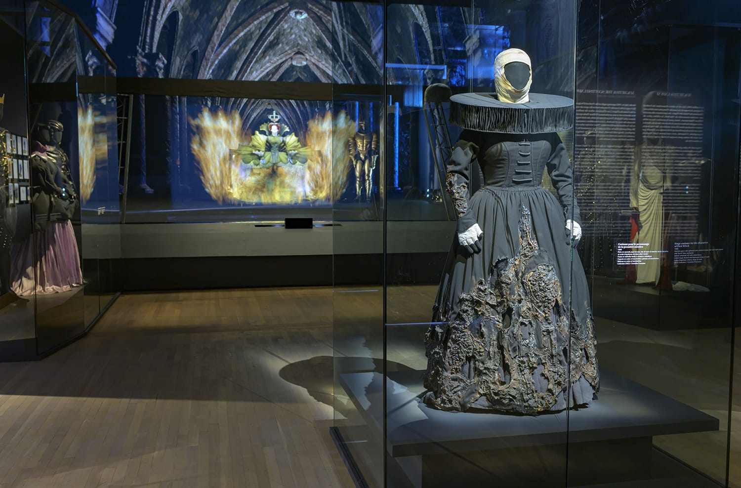 Mannequin wearing an elaborate historical costume in a dimly lit museum exhibit with arched ceiling and visual display in background