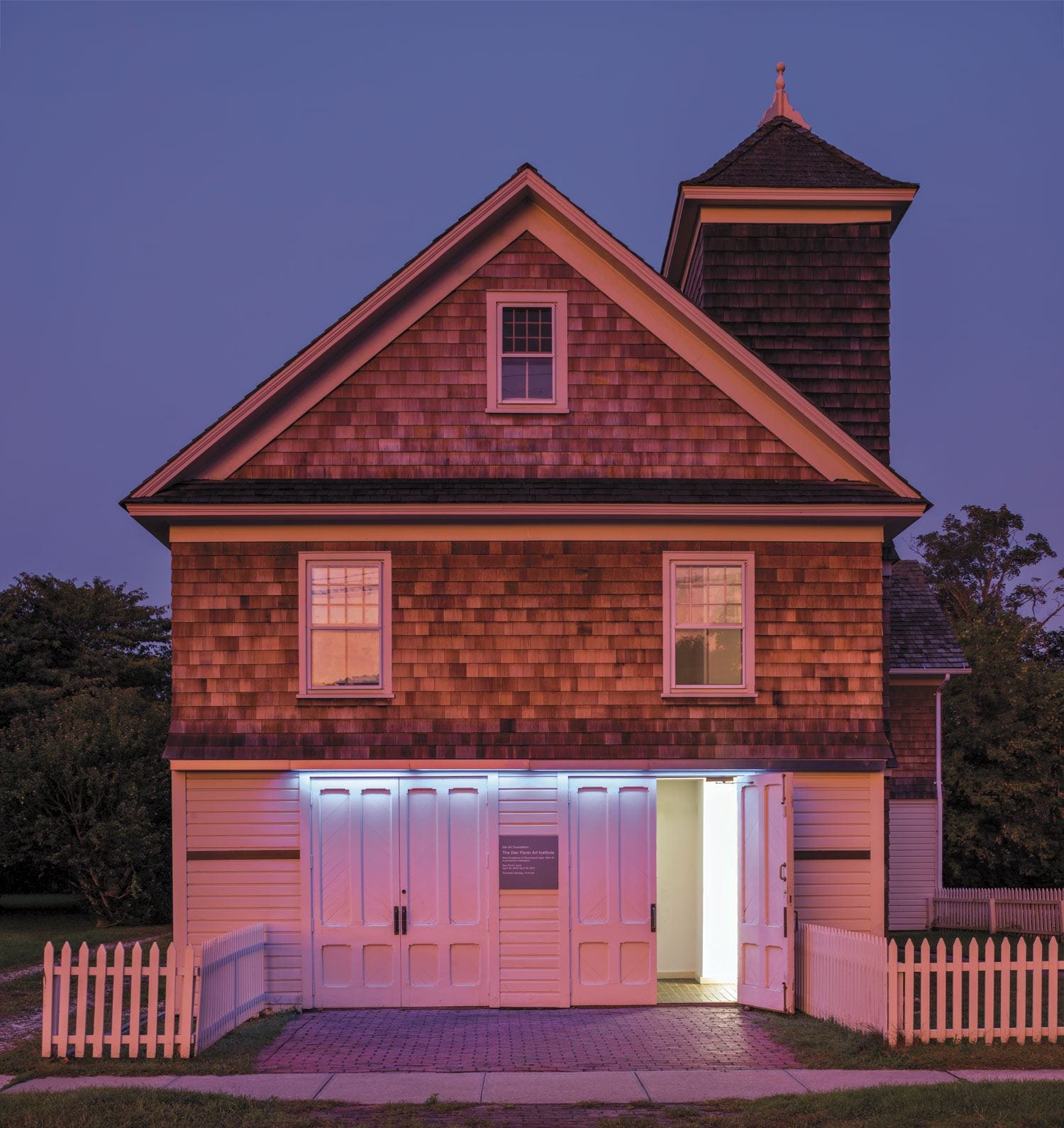 A quaint barn with glowing white doors and trimmed hedges under a dusky purple sky.