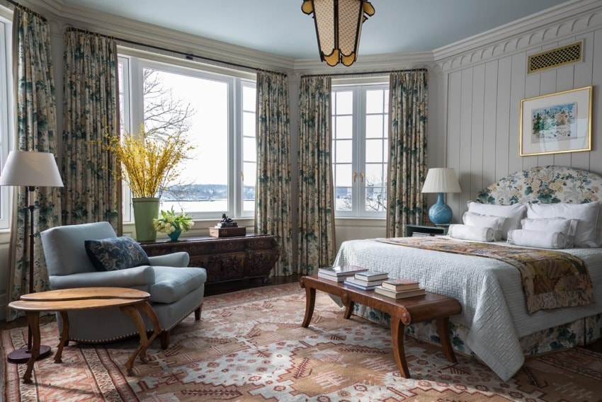 Elegant bedroom with a large window, patterned curtains, a cozy armchair, and a decorated bed, framed artwork on the wall.
