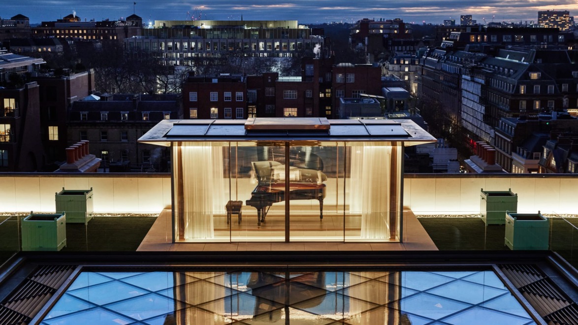 A modern rooftop room with a grand piano, surrounded by cityscape at dusk and a reflective glass surface below.
