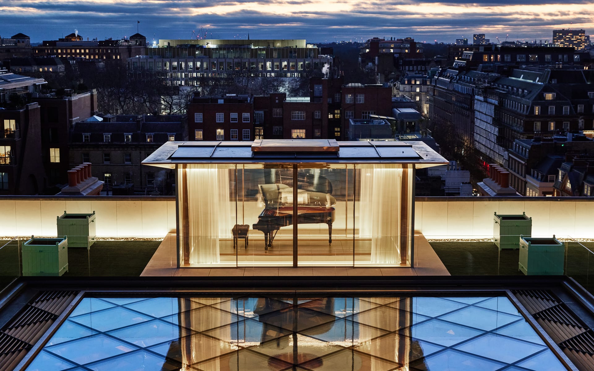 A modern rooftop room with a grand piano, surrounded by cityscape at dusk and a reflective glass surface below.